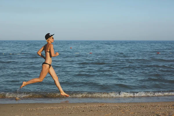 Aktive, sportliche Frau läuft entlang der Meeresbrandung am Wasserpool, um fit und gesund zu bleiben. Sonnenuntergang schwarzer Sandstrand Hintergrund mit Sonne. Frauenfitness, Jogging-Workout und sportliche Betätigung im Sommer-Familienurlaub. — Stockfoto