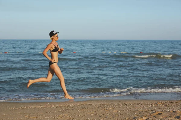 Mulher esportiva ativa correr ao longo do oceano surf por piscina de água para manter a forma e saúde. Pôr do sol areia preta praia fundo com sol. Aptidão da mulher, treino de corrida e atividade esportiva nas férias em família de verão . — Fotografia de Stock