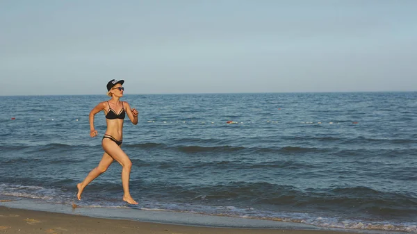 Δραστήρια σπορ γυναίκα τρέχει κατά μήκος του ωκεανού από την πισίνα με νερό για να κρατηθείτε σε φόρμα και υγεία. Με φόντο ηλιοβασίλεμα μαύρη παραλία με ήλιο. Γυναίκα γυμναστήριο, τζόκινγκ προπόνηση και αθλητικών δραστηριοτήτων για οικογενειακές διακοπές καλοκαίρι. — Φωτογραφία Αρχείου
