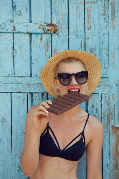 Young attractive woman in sunglasses and in straw hat eating chocolate on a blue background. Film efect — Stock Photo, Image