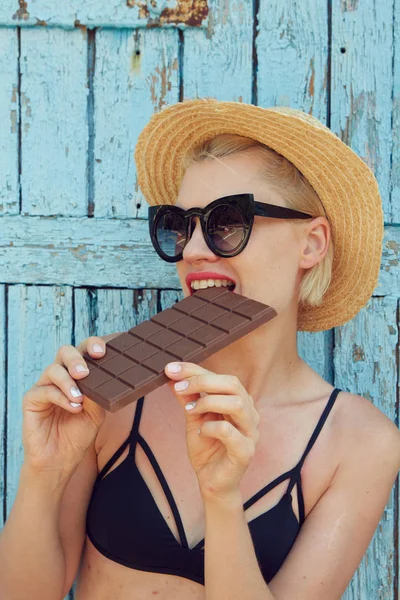 年轻迷人的女人在太阳镜和草帽吃巧克力的蓝色背景。电影影响 — 图库照片