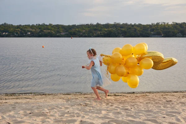 Kleines Mädchen mit vielen goldenen Luftballons am Strand bei Sonnenuntergang — Stockfoto