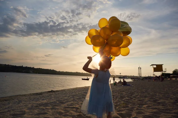 Niña con muchos globos dorados en la playa al atardecer — Foto de Stock