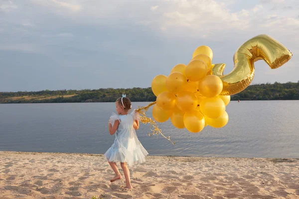 Kleines Mädchen mit vielen goldenen Luftballons am Strand bei Sonnenuntergang — Stockfoto