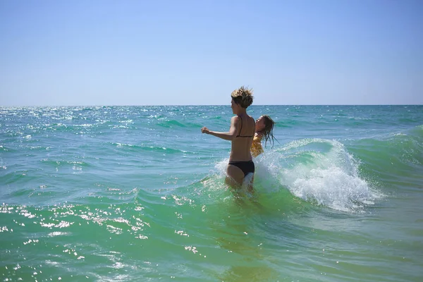 Summer happy family of six years blonde child playing and jumping water waves embracing woman mother in sea shore beach — Stock Photo, Image
