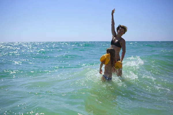 Summer happy family of six years blonde child playing and jumping water waves embracing woman mother in sea shore beach — Stock Photo, Image
