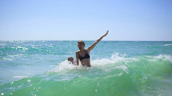 Summer happy family of six years blonde child playing and jumping water waves embracing woman mother in sea shore beach — Stock Photo, Image