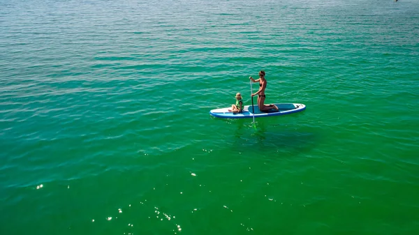 Little girl in a life vest sitting on the paddle board with mother — Stock Photo, Image