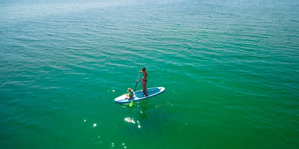 Little girl in a life vest sitting on the paddle board with mother — Stock Photo, Image