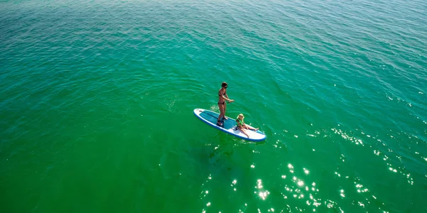 Little girl in a life vest sitting on the paddle board with mother — Stock Photo, Image