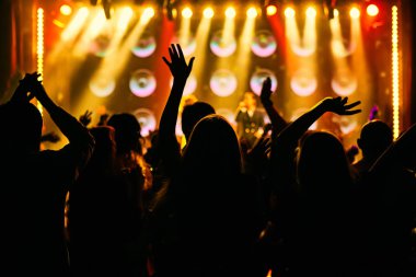 Bir müzik konserinde kalabalık, seyirciler parlak sahne ışıklarının önünde el kaldırıyor..