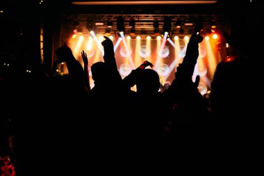 Bir müzik konserinde kalabalık, seyirciler parlak sahne ışıklarının önünde el kaldırıyor..