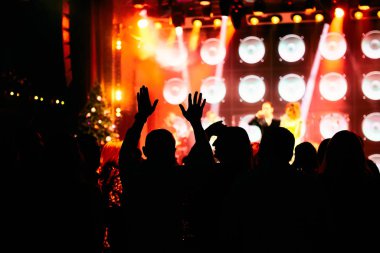 Bir müzik konserinde kalabalık, seyirciler parlak sahne ışıklarının önünde el kaldırıyor..