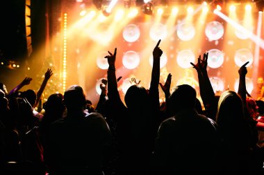 Bir müzik konserinde kalabalık, seyirciler parlak sahne ışıklarının önünde el kaldırıyor..