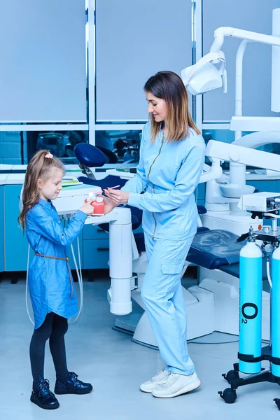 Lasten hammaslääkäri osoittaa perus hampaiden hygienia periaatteet. Naispuolinen pediatrinen hammaslääkäri osoittaa hoitoa suloiselle pikkutytölle sairaalassa. Hammaslääkäri ja lapsi kaapissa. Pieni tyttö... — kuvapankkivalokuva