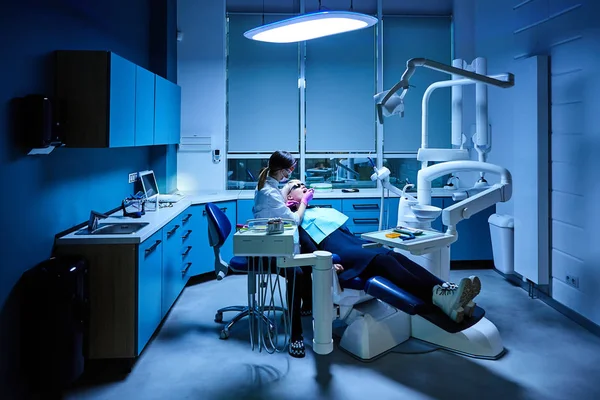 Dentista femenina revisa y repara muy cuidadosamente los dientes de su joven paciente. Mujer joven y feliz sentada en silla dental en el consultorio del dentista —  Fotos de Stock