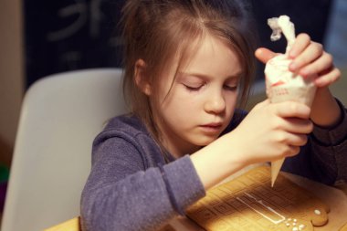 Mutlu küçük kız ev yapımı zencefilli kurabiyeleri ahşap masada jöle kullanarak süslüyor..