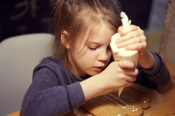 木のテーブルの上に釉薬を使用して自家製のジンジャーブレッドクッキーを飾る幸せな女の子. — ストック写真