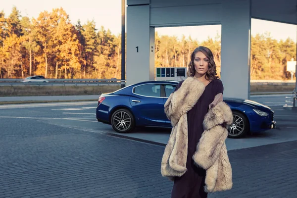 The elegant blonde beautiful woman posing near luxury vehicle. Beautiful young woman with blue luxury car. Sexy female enjoying trip on luxury modern car. Fashionable lifestyle concept.