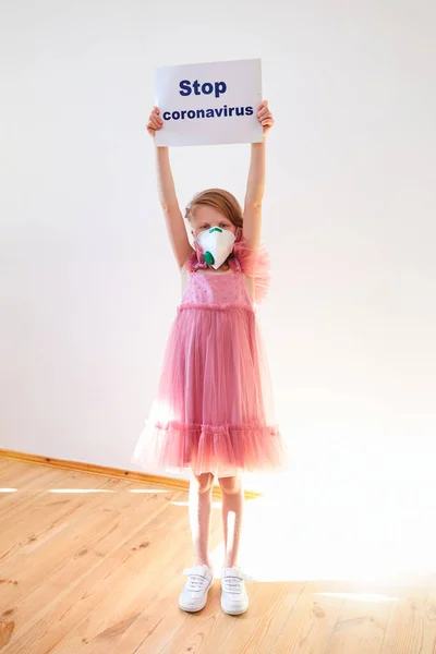 Little girl 7 years old in a medical mask shows banner Stop Coronavirus at home with flecks of sunlight. Concept 2019-nCov. Corona virus outbreaks. Epidemic virus Respiratory Syndrome. Banner for 2019 — Stock Photo, Image