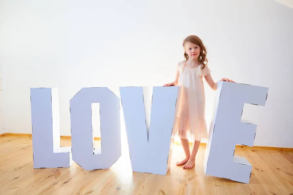 7 살 여자 아이가 큰 판지 3 차원 받침대 글자를 가지고 장난을 치고 있습니다. 밝은 배경에 흰색 판지로 사랑이라는 단어를 만들고 있습니다. 관계 개념. — 스톡 사진