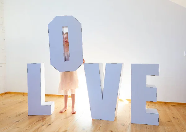 7 살 여자 아이가 큰 판지 3 차원 받침대 글자를 가지고 장난을 치고 있습니다. 밝은 배경에 흰색 판지로 사랑이라는 단어를 만들고 있습니다. 관계 개념. — 스톡 사진