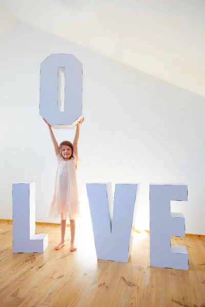 Девочка 7 лет играет с большим картонным 3D стоячие буквы образуя слово ЛЮБОВЬ из белого картона на светлом фоне. Концепция отношений . — стоковое фото