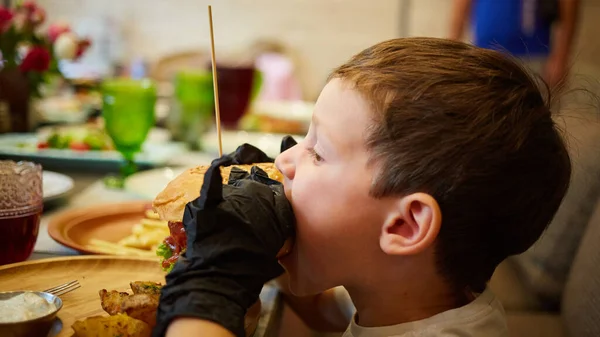 El niño guapo comiendo hamburguesa con guantes negros de goma. — Foto de Stock
