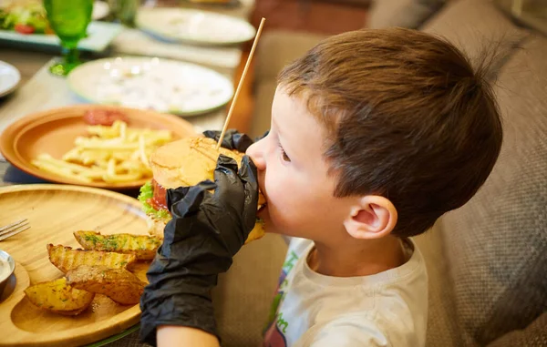 The handsome little boy eating burger in black rubber gloves. — Stock Photo, Image