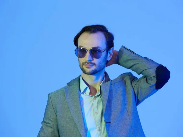 Elegante jonge knappe man met een zonnebril en een stijlvol grijs jasje. Studio modeportret — Stockfoto