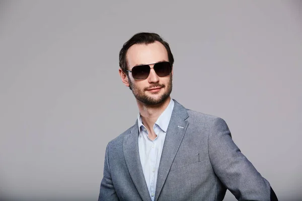 Elegant young handsome man in sunglasses and in stylish gray jacket. Studio fashion portrait. — Stock Photo, Image