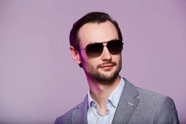 Elegante jonge knappe man met een zonnebril en een stijlvol grijs jasje. Studio modeportret. — Stockfoto