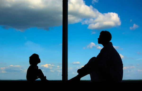 Siluetas de mamá con una hija de 3 años delante de una gran ventana con cielo azul y nubes. Permanecer en casa para la prevención de cuarentena de la pandemia de coronavirus. Autoaislamiento — Foto de Stock