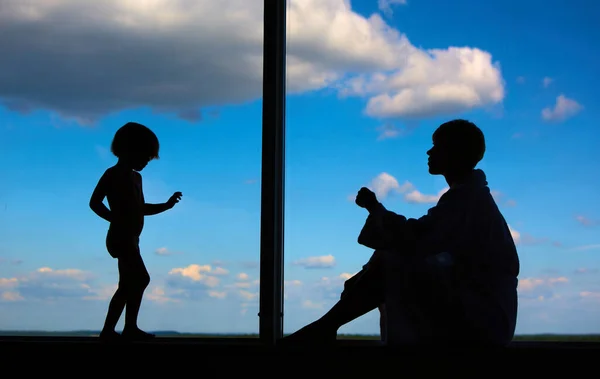 Siluetas de mamá con una hija de 3 años delante de una gran ventana con cielo azul y nubes. Permanecer en casa para la prevención de cuarentena de la pandemia de coronavirus. Autoaislamiento — Foto de Stock