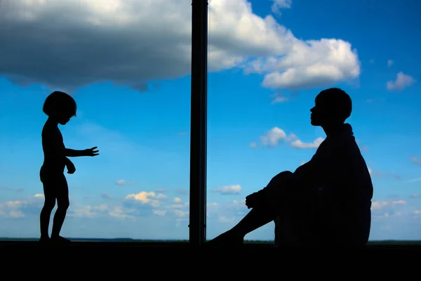 Siluetas de mamá con una hija de 3 años delante de una gran ventana con cielo azul y nubes. Permanecer en casa para la prevención de cuarentena de la pandemia de coronavirus. Autoaislamiento — Foto de Stock