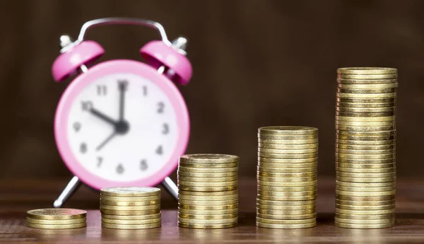 Financial freedom concept. Gold money coins stack with pink alarm clock on brown background.