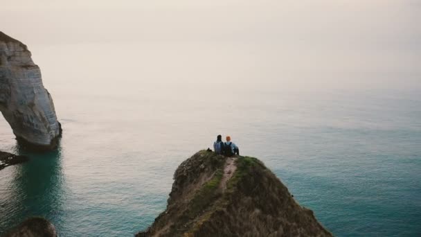 ドローンは幸せなロマンチックなカップルの上に傾き日没の海の景色を見て有名なノルマンディー海岸の崖の上に座って — ストック動画