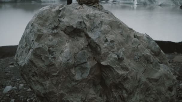 若い男が一人で アイスランドの氷ラグーンで氷河を探して火山岩の上に座っての背面図 — ストック動画
