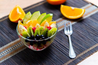 şeffaf cam tabakta meyve salatası. Siyah bir bambu hasırın üzerinde servis edilmiş. Kesilmiş portakalların arka planında. Metal çatallı taze meyve salatası.
