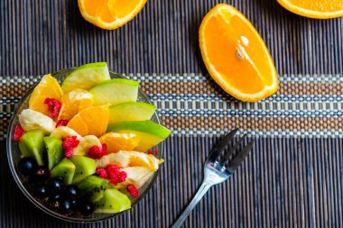 şeffaf cam tabakta meyve salatası. Siyah bir bambu hasırın üzerinde servis edilmiş. Kesilmiş portakalların arka planında. Metal çatallı taze meyve salatası.