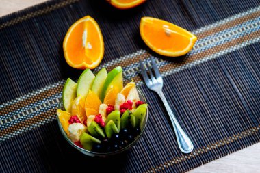şeffaf cam tabakta meyve salatası. Siyah bir bambu hasırın üzerinde servis edilmiş. Kesilmiş portakalların arka planında. Metal çatallı taze meyve salatası.
