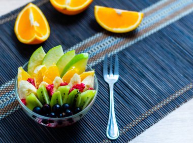 şeffaf cam tabakta meyve salatası. Siyah bir bambu hasırın üzerinde servis edilmiş. Kesilmiş portakalların arka planında. Metal çatallı taze meyve salatası.
