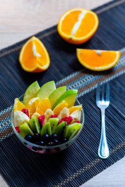 şeffaf cam tabakta meyve salatası. Siyah bir bambu hasırın üzerinde servis edilmiş. Kesilmiş portakalların arka planında. Metal çatallı taze meyve salatası.