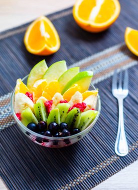 şeffaf cam tabakta meyve salatası. Siyah bir bambu hasırın üzerinde servis edilmiş. Kesilmiş portakalların arka planında. Metal çatallı taze meyve salatası.