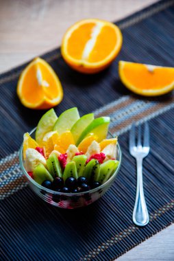 şeffaf cam tabakta meyve salatası. Siyah bir bambu hasırın üzerinde servis edilmiş. Kesilmiş portakalların arka planında. Metal çatallı taze meyve salatası.