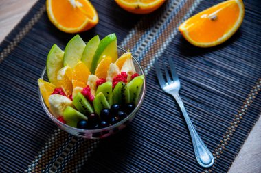 şeffaf cam tabakta meyve salatası. Siyah bir bambu hasırın üzerinde servis edilmiş. Kesilmiş portakalların arka planında. Metal çatallı taze meyve salatası.