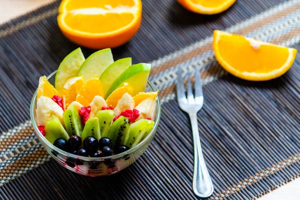 şeffaf cam tabakta meyve salatası. Siyah bir bambu hasırın üzerinde servis edilmiş. Kesilmiş portakalların arka planında. Metal çatallı taze meyve salatası.