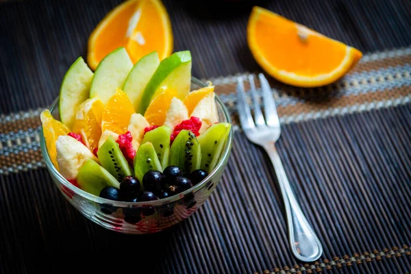 şeffaf cam tabakta meyve salatası. Siyah bir bambu hasırın üzerinde servis edilmiş. Kesilmiş portakalların arka planında. Metal çatallı taze meyve salatası.