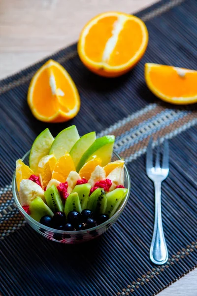 şeffaf cam tabakta meyve salatası. Siyah bir bambu hasırın üzerinde servis edilmiş. Kesilmiş portakalların arka planında. Metal çatallı taze meyve salatası.