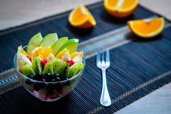 透明ガラス板のフルーツサラダは みじん切りオレンジを背景に濃い竹のマットで供される 金属フォーク付きのクローズアップ新鮮なフルーツサラダ — ストック写真